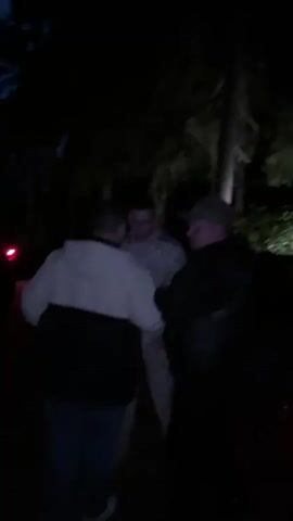 The video shows a group of people outdoors at night in a wooded area. The lighting is dim, with some artificial light sources visible, likely from a vehicle. The camera pans and moves, suggesting people are walking or moving around.

**Subjects:** The primary subjects are several individuals, their faces and features are mostly obscured by the darkness and the camera's movement. One person in the foreground is partially visible and appears to be looking away from the camera. Other individuals are seen in the background, some appearing to be wearing casual clothing, and one person in what looks like camouflage attire. There are trees and foliage indicating a natural, wooded setting. A vehicle with its lights on is briefly visible in the distance.

**Activity/Event:** The people appear to be gathering or moving through the wooded area at night. The context suggests a casual outing, perhaps a walk or a meeting in a secluded outdoor location.

**Notable Details:**
*   **Time of Day:** Night, as indicated by the darkness and the need for artificial lighting.
*   **Weather:** Not directly discernible from the video, but the outdoor setting implies it.
*   **Location Cues:** The presence of dense trees and undergrowth strongly suggests a forest or wooded park. The mention of "Sosnovyi, Russia" provides a specific geographical context.
*   **Emotions:** Due to the low light and obscured faces, it's difficult to discern specific emotions, but the general atmosphere appears relaxed and informal.
*   **Visible Text:** No discernible text is visible in the video.