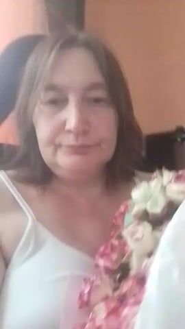In this media file, a woman with shoulder-length brown hair, wearing a white camisole, is holding a bouquet of pink and white roses. She is positioned in front of an orange wall, with a dark object, possibly a chair, visible behind her. The lighting suggests it is daytime. The woman's expression is neutral, and she appears to be in a relaxed indoor setting. The activity appears to be a casual presentation of the flowers. There is no visible text in the media file. The location context is Berezynka, Ukraine.