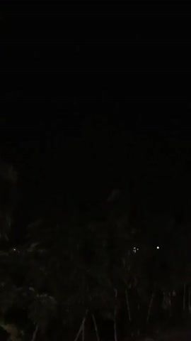 This is a very dark video. It appears to be a recording of an outdoor scene at night. The bottom portion of the frame shows the silhouettes of trees or bushes, with some sparse vegetation visible. In the middle ground, there are a few faint, bright specks of light, possibly from distant artificial lights or reflections. Towards the right side of the frame, a brighter, rectangular light source is visible, suggesting a building or structure with illuminated windows. The upper portion of the frame is completely black, indicating a dark sky with no visible stars or moon. The camera is stationary, and no movement or action is discernible.