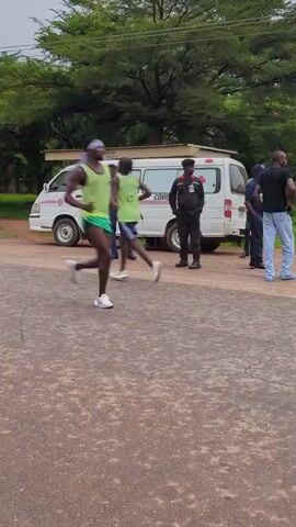 Two young men in athletic wear are running on an asphalt road, with green and white uniforms and visible numbers on their backs. The runner in the lead wears number 15. They run past a white van and a man who appears to be a security guard standing nearby. Trees line the sides of the road. The video follows the runners as they continue down the road, passing another man standing on the side. Overhead power lines are visible against the sky.
