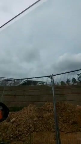 The video shows a construction site with a skeletal building frame and piles of dirt in the foreground. A metal fence encloses the site. The sky is overcast with clouds. The view is from inside a vehicle, with a side mirror visible on the left side of the frame. The footage is shaky, suggesting movement.
