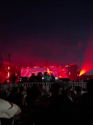 The video shows a crowd of people gathered at an outdoor event at night. A stage is set up in the background with bright red lights illuminating a screen displaying abstract visuals. Several people, presumably performers or DJs, are visible on the stage behind a railing. The crowd is mostly silhouetted against the stage lights, but some individuals in the foreground are more clearly visible. One person in the foreground is holding up a drink, their face partially obscuring the view. The overall atmosphere appears to be that of a concert or music festival.