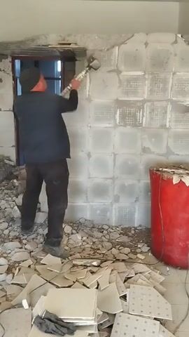 Here is a description of the video:

A man is seen demolishing a wall in a room. He is wearing a dark beanie, a dark vest over a long-sleeved shirt, and dark pants. The man is wielding a large hammer, striking the wall repeatedly. The wall is made of square concrete blocks. Debris from the wall, including broken tiles and chunks of concrete, is scattered on the floor around him. To the right of the man is a tall, red barrel. A doorway, framed in dark wood, is visible through a hole in the wall. The overall scene suggests a construction or demolition project is underway.