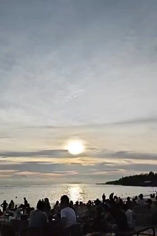 The video shows a beach at sunset. The sky is cloudy, with the sun shining brightly through a gap in the clouds. The sea is calm and reflects the sunlight. In the foreground, there are many people sitting and standing, likely at a beachside gathering or restaurant. Their figures are mostly silhouetted against the bright sky. The overall atmosphere appears relaxed and enjoyable.