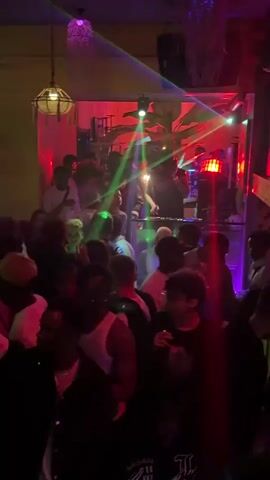 The video captures a lively scene inside a nightclub or bar, likely in Shenyang City, China, at night. A diverse crowd of people is seen dancing, moving to the music, and socializing in a dimly lit, energetic environment.

The focal point is a DJ booth where a person is operating DJ equipment, including turntables and a mixer, appearing to be actively performing for the crowd. Above the DJ, and throughout the venue, vibrant club lighting consisting of red, green, and blue laser beams and spotlights sweeps across the room, creating dynamic visual effects. Colored overhead lamps, some appearing to be woven or textured, contribute to the atmospheric lighting.

The decor features large, tropical-looking artificial plants, adding a themed ambiance to the venue. People in the crowd are seen holding drinks and engaging with the music. The overall mood is energetic and festive, with individuals appearing to be enjoying the music and the atmosphere. No visible text is present in the video.