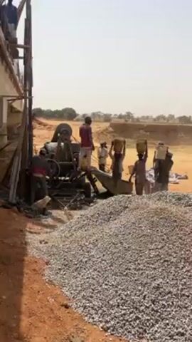 The video shows a construction site in a dry, dusty environment. In the foreground, there's a large pile of gravel next to a red dirt embankment. Several workers are visible, operating machinery and carrying buckets. One worker is tending a cement mixer, while others carry buckets on their heads, presumably filled with materials for construction. A partially constructed concrete bridge spans the scene, supported by scaffolding. The sky is overcast and the background reveals a flat, arid landscape.