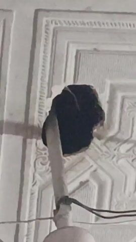The video shows a close-up view of a ceiling fan installation, focusing on the point where the fan's downrod meets the ceiling. The ceiling is white with a decorative, patterned design. A black, possibly damaged or improperly installed, ceiling box is visible, partially concealed by the ceiling's design. Wires are seen extending from the downrod and into the electrical box.