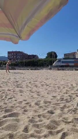The video captures a bright, sunny scene at a sandy beach in Premià de Mar, Spain. It opens with a view from under a striped beach umbrella. A person, a female wearing a bikini, walks on the sand on the left side of the frame. In the background, a train approaches on tracks running parallel to the beach, with buildings visible behind the tracks.

As the video progresses, the camera zooms in, revealing a black and white dog, resembling a Border Collie, running excitedly on the sand. The dog moves from left to right across the frame, kicking up sand as it goes. A white, blue, and orange commuter train passes quickly on the tracks. The word "Renfe" is clearly visible on the side of the train cars. Further along the train, the text "Rodalies" and its distinctive "R" logo are also visible. The dog appears to be reacting to or playfully chasing the passing train, briefly looking back at it. The sky is clear and blue. Some graffiti is visible on the train carriages.