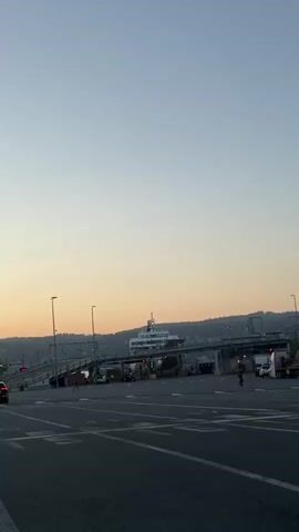 The video shows a ferry terminal with multiple ferries docked and vehicles lined up, including semi-trucks, awaiting transport. The sky is a pale blue to orange gradient, indicating either sunrise or sunset. A few pedestrians are visible, walking across the road in front of the camera. The location appears to be a busy transportation hub.