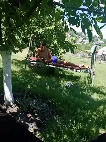 A young man is lying on a raised cot in a grassy area under a tree. He is shirtless and wearing blue shorts. He is holding a phone and appears to be looking at it. The cot has a patterned mattress and is supported by metal legs. The background shows more trees, vegetation, and some buildings in the distance. The lighting suggests it is daytime, likely sunny.