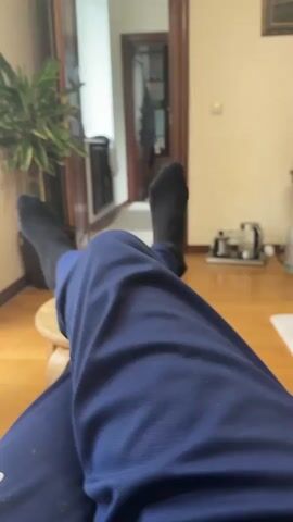 The video shows a person sitting with their legs stretched out and feet resting on a small wooden stool. They are wearing blue sweatpants and black socks. The scene takes place in a room with wooden floors. A potted plant is visible to the left, and a doorway is behind the person. A small machine and a white mat are also on the floor in the background.