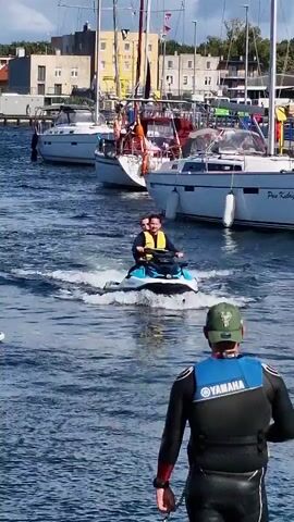 A jet ski with two individuals wearing life vests travels away from the camera on choppy blue water. The person in the front is driving, and the person behind them is looking ahead. Both appear to be smiling. Behind the jet ski, several sailboats are docked. In the foreground, a person wearing a black wetsuit with a blue and white Yamaha vest is facing away from the camera, looking towards the jet ski. They are wearing a green cap. The water is calm except for the wake of the jet ski. Buildings and trees are visible in the background beyond the docked boats.