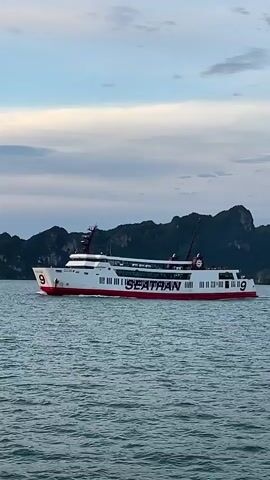 A ferry with the name SEATRAN and the number 9 on it sails on the water. The ferry is white with a red stripe along the bottom and red trim. There are several windows along the side of the ferry. In the background, there are green islands with dark green trees on them. The sky is blue with some white clouds. The water is choppy.