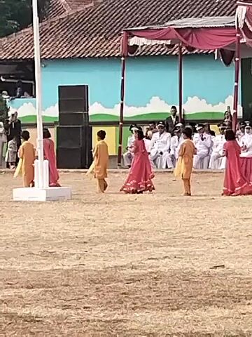 A group of children, some wearing orange outfits and others pink dresses, are performing a dance on a dry, grassy field in front of a building with a red-tiled roof and a blue mural. A white flagpole stands to the left, with large black speakers behind it. In the background, adults sit on chairs under a red draped canopy. The children are in various poses, some facing each other, others turning, and one child in orange appears to be reaching out to a child in pink. The overall impression is that of a school or community event, possibly a cultural performance.
