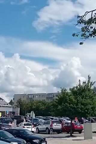The video captures an outdoor scene with a sky filled with white clouds and a blue sky. In the foreground, a tree branch with green leaves is visible. Below the sky, there is a parking lot with several cars, including white, red, and black vehicles. A large building with multiple windows and a gray facade is situated behind the parking lot. To the right, a modern building with large glass windows and concrete pillars is shown. There are also some black signboards hanging from the building. Another building with a lighter colored facade and windows is visible in the background to the right. A person on a bicycle is seen riding on a paved area in front of some modern structures and vegetation. The scene appears to be a public space, possibly a commercial area or an urban environment.
