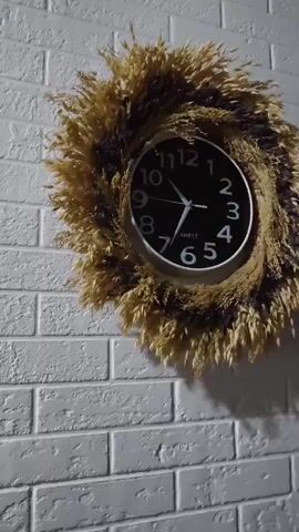 A close-up video shows a clock on a white brick wall. The clock has a black face with white numbers and hands. It is surrounded by a decorative wreath made of dried grasses or reeds. The video pans slightly, showing the texture of the brick wall and the details of the clock and wreath. The lighting is dim, casting a shadow of the wreath on the wall.