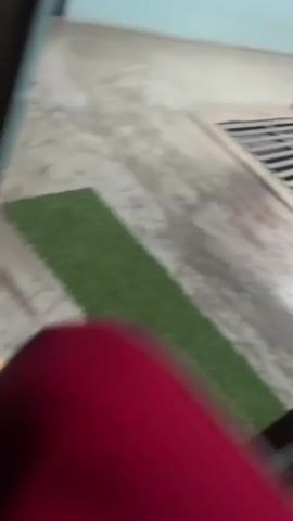 A handheld, shaky video shows a close-up view of a dark, striped fabric draped over a long, narrow metal grate. The fabric is dark brown with thin red and white stripes. Behind the fabric, the grate appears to be set into a concrete surface. The camera then tilts upwards to reveal a skylight with horizontal slats allowing sunlight to pass through. The overall environment appears to be an indoor space with concrete flooring and light blue walls. The video is brief and lacks any discernible action or interaction.