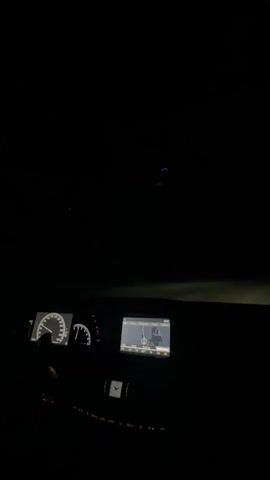 The video captures a first-person perspective from the inside of a car driving at night in what is likely Golemo Selo, Bulgaria, given the context. The dashboard is illuminated, featuring the speedometer and RPM gauges on the left, and a central infotainment screen displaying a GPS navigation map. Below the screen, an analog clock and various climate control buttons are visible. A driver's hand is partially visible on the steering wheel. Outside the windshield, the surroundings are dark, but the bright headlights of an oncoming vehicle briefly illuminate the road ahead as it passes. The overall scene is calm, with no specific weather events apparent. Electronic music with a strong beat plays in the background throughout the video.
