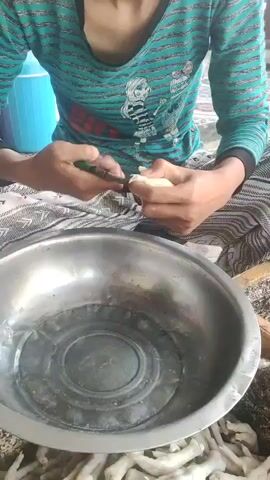 A person wearing a turquoise and black striped shirt with a graphic print sits on the ground, preparing food. They are using a small knife to trim a piece of white material, likely food, held in their left hand. In front of them is a large, shallow metal bowl containing clear liquid and a circular insert. Below the bowl, a large pile of what appears to be chicken feet is visible. The background shows a blue container and other indistinct objects. The lighting is bright, suggesting an outdoor or well-lit indoor environment.