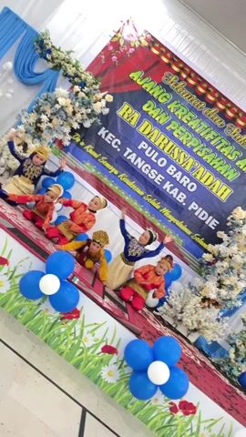 A group of six young children in traditional Indonesian clothing are performing a dance on a stage. The children, appearing to be of primary school age, are dressed in colorful costumes featuring gold, red, and blue. Some wear headdresses. They are arranged in two rows on a patterned rug. The stage is decorated with blue and white fabric draped at the top, floral arrangements, and clusters of blue and white balloons. A large banner in the background reads in Indonesian: "Selamat dan Sukses - AJANG KREATIFITAS DAN PERPISAHAN RA DARUSSAYADAH KEC. TANGLSE KAB. PIDIE". This translates to "Congratulations and Success - Creativity and Farewell Event RA Darussayadah Tangse District, Pidie Regency". The lighting suggests an indoor event. The video is captured from a slightly elevated angle and tilted.