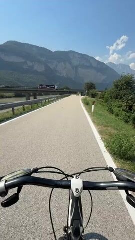 The video shows a first-person perspective of someone riding a bicycle on a paved path. The path runs alongside a river, with a bridge crossing it in the distance. Mountains with rocky faces and patches of green vegetation form the backdrop. The sky is a clear blue with a few scattered white clouds. The handlebars and front wheel of the bicycle are visible in the foreground. A white line marks the edge of the bicycle path. The camera is tilted slightly upwards, emphasizing the expansive sky and mountains.