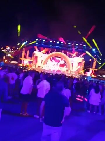 The video shows a crowded outdoor concert at night. A large stage is lit up with colorful lights and lasers.  The stage has a large heart decoration. People are standing and watching the performance. There is a bar with "ABSOLUT." signage in the foreground. The sky is dark, and there are some buildings visible in the distance. The overall atmosphere is energetic and festive.