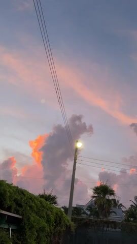 The video shows a slow pan of a sunset sky with dramatic clouds. The clouds are illuminated with vibrant orange and pink hues from the setting sun, contrasting with the darkening blue sky. Silhouetted against the sky are the tops of palm trees and some buildings. A utility pole with wires and a streetlamp is visible on the left side of the frame, and another streetlamp is visible on the right side. The overall impression is one of a beautiful and peaceful evening scene.