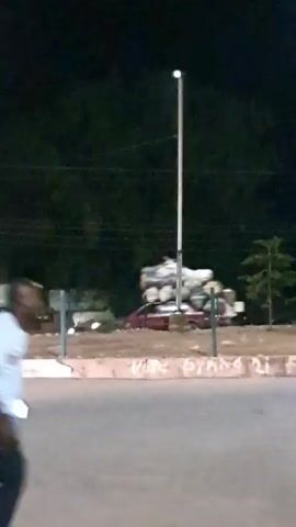 A blurry nighttime video shows a dark-colored car driving down a road. The car is heavily laden with a large stack of logs on its roof. A person in a white shirt and dark pants is visible in the foreground, blurred as they move. Other blurred figures and structures are visible in the background, suggesting an urban or semi-urban environment. The lighting is dim, with some streetlights or building lights visible.