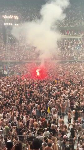 This media file depicts a large crowd of people gathered in a stadium-like setting, possibly a concert or sporting event, given the tiered seating visible in the background. A prominent red flare is lit in the midst of the crowd, emitting a thick cloud of white smoke that billows upwards. The overall atmosphere appears to be energetic and enthusiastic, with many individuals raising their arms. The time of day is difficult to ascertain definitively, but the lighting suggests it is likely evening or indoors. The presence of a flare indicates a potentially passionate or celebratory moment. There is no visible text in the media file.