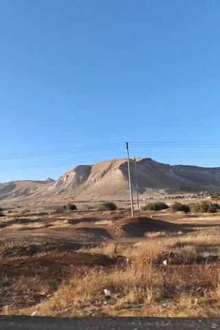 The video shows a dry, arid landscape under a clear blue sky. In the foreground, there is a dirt road with dry grass and brush. In the mid-ground, there are more dry plants and some scattered trees. In the background, there is a large, light-colored mountain with a flat top and steep, rocky sides. There are power lines running across the landscape. In the first few seconds of the video, an Israeli flag on a pole is visible on the right side of the screen. The camera appears to be moving, as the landscape shifts slightly throughout the clip.