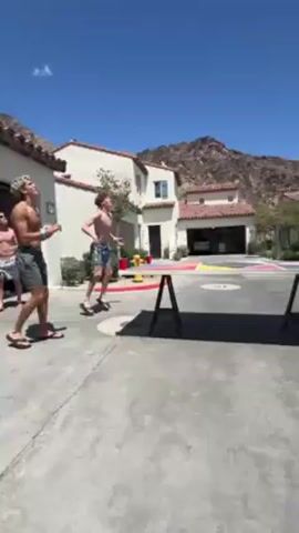 Here is a detailed description of the video:

**Scene:** An outdoor setting, possibly a driveway or parking area, on a bright, sunny day. In the background are buildings with a Spanish-style architecture. A large mountain is visible in the distance.

**People:** The video features multiple young men, mostly shirtless and wearing shorts and flip-flops. Some are actively engaged in a game, while others are spectating or observing.

**Objects:** Key objects include two tables set up with red cups at each end, suggesting a beer pong or similar game setup. There are also cans/bottles of beverages, possibly alcoholic, near the tables. A decorative chair is visible.

**Actions:** The primary action is the playing of a game similar to beer pong. Participants are seen throwing ping pong balls towards the cups, interacting with each other, and celebrating successes.

**Interactions:** The men interact with each other by throwing ping pong balls at the cups.

**Environment:** The setting is a modern-looking outdoor space, with desert plants and mountain views. The location appears to be a residential or resort area, possibly in a desert climate.