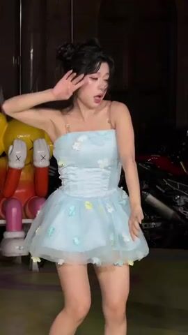 A young Asian woman performs a lively dance routine in what appears to be an urban-themed indoor or covered outdoor space, likely in Nan'an District, China. She is dressed in a light blue strapless mini-dress with a corset-style bodice and a fluffy tulle skirt adorned with small white, yellow, and light blue flower appliqués. Clear shoulder straps are visible, and she wears white socks with light-colored shoes. Her dark hair is styled in two small buns or half-up pigtails, with some loose strands framing her face. She has noticeable makeup and displays a cheerful, confident expression throughout her performance, frequently smiling and engaging with the camera.

The background is dimly lit, suggesting an evening or nighttime setting, with spotlights illuminating the subject. To the left, a large, iconic yellow and red KAWS Companion art figure is seated on a bench. To the right, several motorcycles are parked, including a prominent red and black one. The ground features colorful circular patterns or graffiti. The combination of the art figure and motorcycles creates a modern, artistic, and somewhat edgy atmosphere. The woman's movements are fluid and energetic, involving various poses, arm gestures, and body swaying as she executes her choreography. No visible text is legible in the scene.