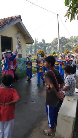 This media file captures a lively scene likely in Posong, Indonesia. The primary subjects are a group of young people, possibly a marching band or a school group, dressed in vibrant blue and yellow uniforms with matching hats. They appear to be in formation, standing in neat rows on a paved outdoor area.

In the foreground, two boys are prominent. One is wearing a red t-shirt and white pants, gesturing with his right hand. Behind him, another boy in a brown t-shirt with a red strap slung across his chest is looking down, seemingly engaged with something he's holding. To the left, a smaller building with a tiled roof is visible. A mural adorns its exterior wall, depicting stylized figures, possibly relating to a local story or tradition.

The background suggests an outdoor setting with greenery and trees, indicating it's likely daytime. The bright lighting implies clear or partly cloudy weather. The overall atmosphere seems energetic and possibly preparatory for a performance or parade, given the uniforms and organized arrangement of the individuals. The presence of other people in the background suggests a public event or gathering. There is no visible text in the media.
