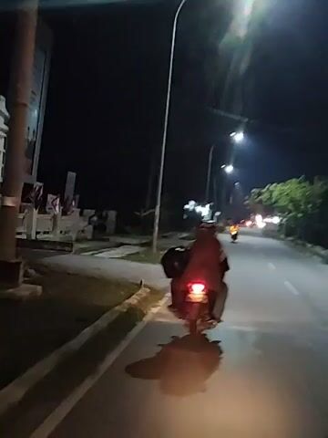 The video captures a night-time scene of a person riding a motorcycle on a road. The rider, covered in a brown garment, is seen from behind, with their motorcycle's taillight illuminated. The road is paved and appears to have streetlights lining one side. Trees are visible in the background, along with some blurry structures and distant lights. The motorcycle carries luggage on the back. The perspective suggests the video is being filmed from another moving vehicle or by a passenger on the same motorcycle, looking back. The lighting conditions are low, with the primary illumination coming from the streetlights and the motorcycle's taillight, casting shadows on the road. The overall ambiance is one of travel in darkness.