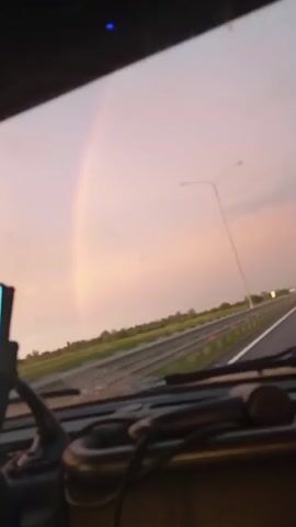 The video shows a view from inside a vehicle, looking out through the windshield. A faint rainbow is visible in the sky to the left. The sky is a pale pinkish-purple color. A highway stretches into the distance with a line of streetlights along the right side. Trees and fields line the sides of the highway. The dashboard of the vehicle is visible in the foreground, with some dark objects on it. The footage is grainy and appears to have been recorded with a low-resolution camera.