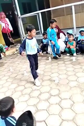 The video shows a group of young children participating in an outdoor game. Two boys are actively playing in the foreground, one in a blue and black tracksuit and the other in a light blue and white tracksuit. They appear to be navigating around small white cups placed on a hexagonal tiled surface. Several other children, some wearing hijabs and matching uniforms, are seated on a raised platform in the background, watching the game. The environment suggests a school or community setting, with a building visible behind the seated children and some greenery. The overall atmosphere seems to be one of friendly competition and amusement.