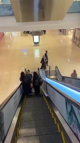The video shows a downward-facing perspective of an escalator in a mall. Several people are visible on the escalator and in the mall area below. On the escalator, there are at least five people descending, with a group of three women in the foreground on the left, followed by two more individuals. To the right on the ascending escalator, there are at least two people. In the mall area, a man in a black shirt and jeans walks away from the escalator, and another man wearing a black robe with white trim walks towards the ascending escalator. Further in the background, several other individuals are visible, some walking and some seated. A digital display screen is also visible in the middle of the mall. The mall floor appears to be polished tile, and the walls are decorated with signage, including one that advertises a "SEASON END SALE UP TO 60% OFF". The lighting is bright, indicating an indoor shopping environment.