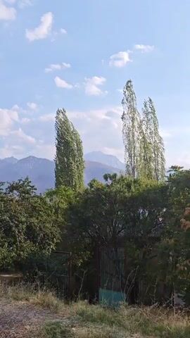 The video pans across a rural landscape under a bright blue sky with scattered clouds. In the foreground, there is a dirt path with dry grass and some sparse vegetation. A rustic fence runs along the edge of the path. Behind the fence, there are trees and bushes, some of which are tall and slender like poplar trees, while others are more dense and leafy. In the middle ground, a small, simple building with a red tiled roof and white walls is visible. Further in the distance, rolling hills and mountains create a layered backdrop. The overall impression is one of a quiet, natural setting in a hilly or mountainous region.