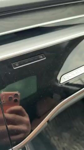 The video shows the interior dashboard of a car, specifically focusing on the "Quattro" emblem. The dashboard is a glossy black finish with silver accents. The camera zooms in slightly on the emblem, then pans upwards along the dashboard showing the silver trim and the wood grain texture.  The reflection of a person holding a cell phone can be seen in the glossy surface.  The outdoor environment is visible through the car's windows.