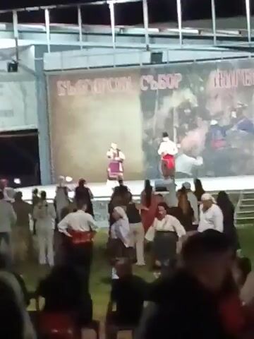 The video captures an outdoor night scene of a cultural event. A brightly lit stage features two individuals in traditional attire. One person stands centrally, holding a microphone and wearing a purple and white folk costume, appearing to perform. To their right, another individual stands wearing a white shirt, red sash, and dark cap. A large banner behind them displays "БЪЛГАРСКИ СЪБОР" (Bulgarian Gathering) along with illustrated scenes. In the foreground, a diverse crowd is gathered on a grassy area, many also dressed in traditional Bulgarian folk costumes, including white shirts with red sashes and patterned dresses. The attendees are standing, observing the stage, and some are subtly moving or swaying.