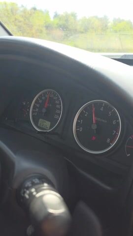 The video shows a driver's perspective from inside a car moving on a highway. The dashboard is visible, showing a speedometer reading around 100 km/h and a tachometer reading around 1000 rpm. The windshield shows the highway ahead, with other cars and a large truck in the distance. The sky is bright, and there are trees alongside the highway. The general impression is of someone driving on a highway during a sunny day.