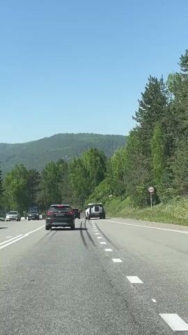 A low-angle shot captures a car traveling along a winding road, with two vehicles ahead in the distance. The car in front on the left is a dark SUV. The vehicle on the right is a white vehicle with a flat rear end, which could be a truck or a specialized vehicle. The road has a solid white line on the left, a broken white line in the middle, and a solid white line on the right. Skid marks are visible on the road surface. The road curves to the left. The scene is set in a natural environment with a steep, green-forested hill on the right and rolling green hills in the distance. The sky is clear and blue.