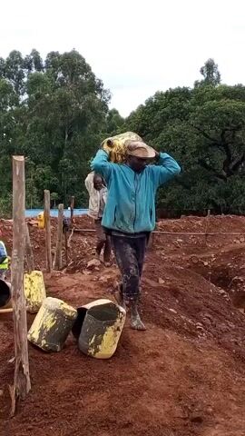 The video shows three men working at what appears to be a construction site. The ground is covered in reddish-brown dirt and mud. Two men are carrying yellow containers, likely filled with something heavy, on their shoulders, while the third man, wearing a blue t-shirt, walks to the side of them. They are surrounded by partially emptied yellow containers and wooden stakes delineating the site.  There are trees and foliage in the background under an overcast sky.
