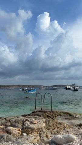 A vertical shot captures a rocky shoreline in the foreground, with the ocean and a fleet of boats in the midground and background. The sky is a mixture of blue and white clouds. In the foreground, there are rocks and pebbles. A metal ladder extends from the shore into the clear, turquoise water. Several boats, including speedboats and a larger white vessel, are anchored in the calm sea. The horizon is visible in the distance, with land and more boats. The scene is serene and offers a sense of a calm day by the water.