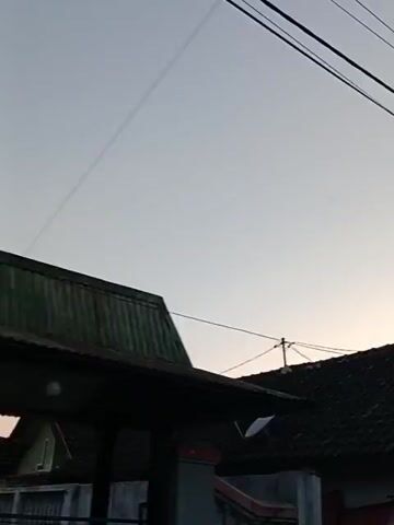 The video pans across a street, showing houses and buildings. The sky is clear and light blue, suggesting it is either sunrise or sunset. The camera begins by focusing on the roof of a building with a dark tiled roof. Red flags are visible hanging on the side of one building. The camera then moves to show a light green two-story building with a balcony. Next, a cream-colored building with a dark roof and a row of windows is shown. Finally, the video ends with a shot of a building with a black metal awning. Power lines are visible throughout the video, crisscrossing the sky.