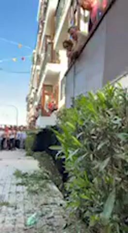 The video shows a street scene in Vejer de la Frontera, Spain, during the daytime with a clear blue sky. The camera is positioned at street level, looking upwards at a multi-story building with balconies.

On the balconies, numerous people are visible, leaning over the railings and looking down, suggesting they are observing an event happening on the street below. More people can be seen on the street level in the background, though they are out of focus. In the foreground, there are dense green bushes or plants, with some trimmed foliage or debris scattered on the ground. A string of colorful flags or bunting is visible stretched across the street in the upper left corner.

An audible voice, likely from a speaker or announcer, is heard in Spanish, mentioning words such as "retirada," "demanda," "animal," and "colaborar," suggesting an organized public event or procession. General crowd noise and murmuring can also be heard. No explicit text is visible in the video.