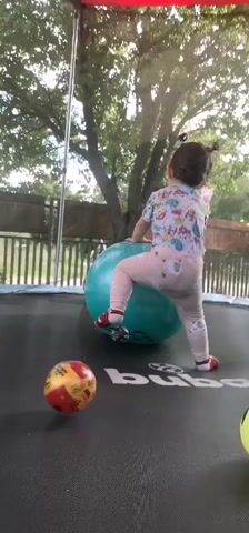 A young child is playing with large inflatable balls and smaller balls on a trampoline. The child is wearing a colorful t-shirt, pink pants, and red shoes. There are several balls on the trampoline, including a large blue inflatable ball with a cartoon owl face, a yellow ball with heart-shaped eyes, and other smaller colored balls. The child is actively interacting with the balls, pushing and bouncing them around. The environment appears to be an outdoor backyard with trees and a fence visible in the background. The trampoline has the "buba" logo on it. The footage is shaky and appears to be recorded by someone holding a camera.