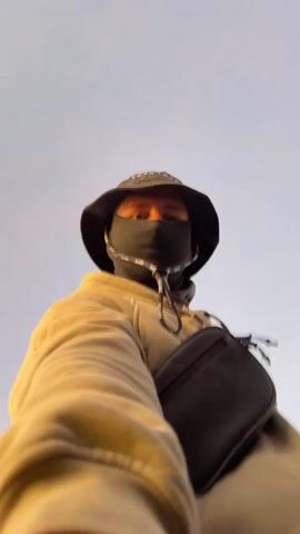 This media file features a person wearing a tan hoodie, a black bucket hat with "BORN TO BE" printed on it, and a black face mask. They are standing on a hillside overlooking a volcanic landscape at sunrise. The sky is painted with soft hues of orange, pink, and blue. Smoke billows from a nearby volcano, creating a dramatic contrast against the serene dawn. The person is holding a camera, taking a selfie with the breathtaking scenery in the background. The overall mood is one of awe and adventure, capturing the essence of exploration and the beauty of nature. The location appears to be Cemorolawang, Indonesia, known for its stunning views of Mount Bromo and its surrounding volcanic craters.