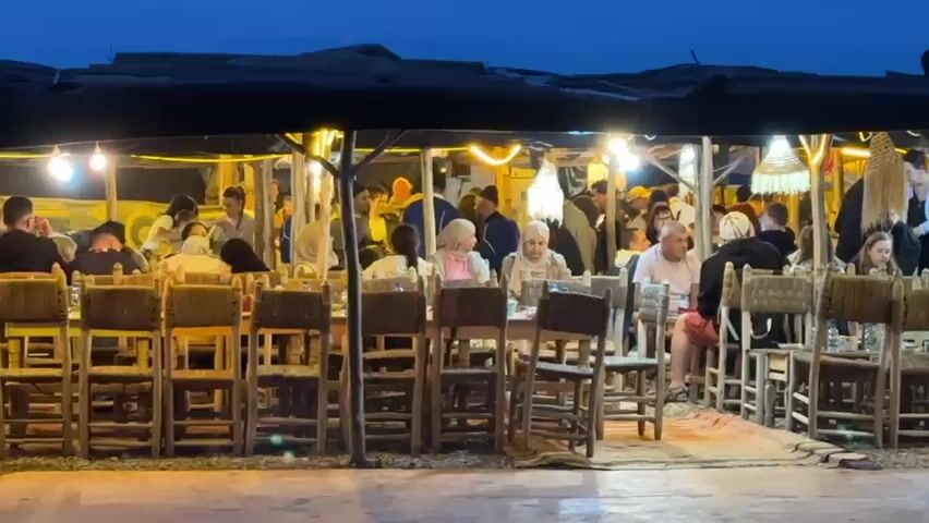 The video captures a lively outdoor dining scene at night, likely in Sidi Badhaj, Morocco. Numerous people are gathered under a large tent or canopy structure, illuminated by warm string lights and decorative lanterns. They are seated at long wooden tables with matching chairs, engaged in conversation, eating, and drinking. Some individuals are seen standing and moving around the bustling area.

The sky is a deep blue, indicating the late evening or early night. The atmosphere appears social and relaxed, with people enjoying their meals and company. The setting evokes a traditional Moroccan market or open-air restaurant. The background is filled with the murmur of conversations and what sounds like traditional music, contributing to the vibrant ambiance. There is no visible text in the frame.