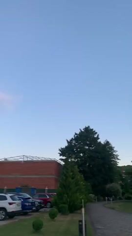 The video shows an outdoor scene with a clear blue sky. In the foreground, there is a paved path curving to the left, bordered by grass. Several trees are visible, some with dense foliage and others more sparse. In the background, there appear to be buildings and a sports field with two goals. The lighting suggests it is either early morning or late evening. There are a few people walking on the path, and some objects that look like air conditioning units are attached to a building. The camera then pans to a building with a glass elevator shaft. The video ends with a view of a sunset with scattered clouds, silhouetted trees, and a car parked on a driveway.